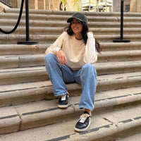 Mujer sentada en unas escaleras exteriores, sonriendo y llevando la gorra KUMI verde oliva junto a una sudadera color natural y vaqueros