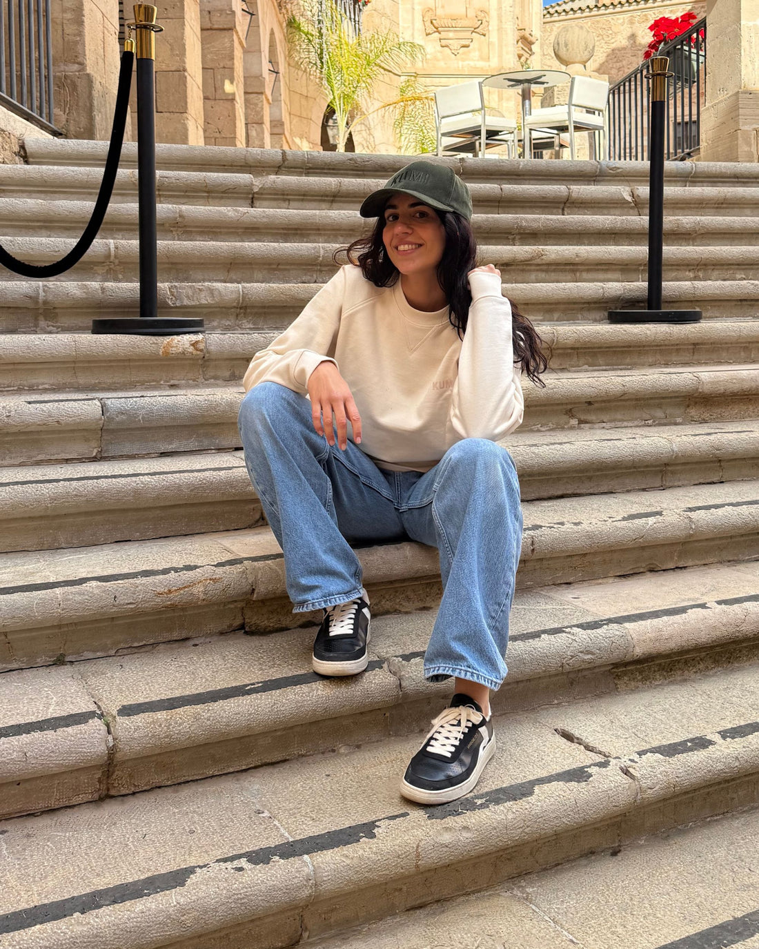 Mujer sentada en unas escaleras exteriores, sonriendo y llevando la gorra KUMI verde oliva junto a una sudadera color natural y vaqueros
