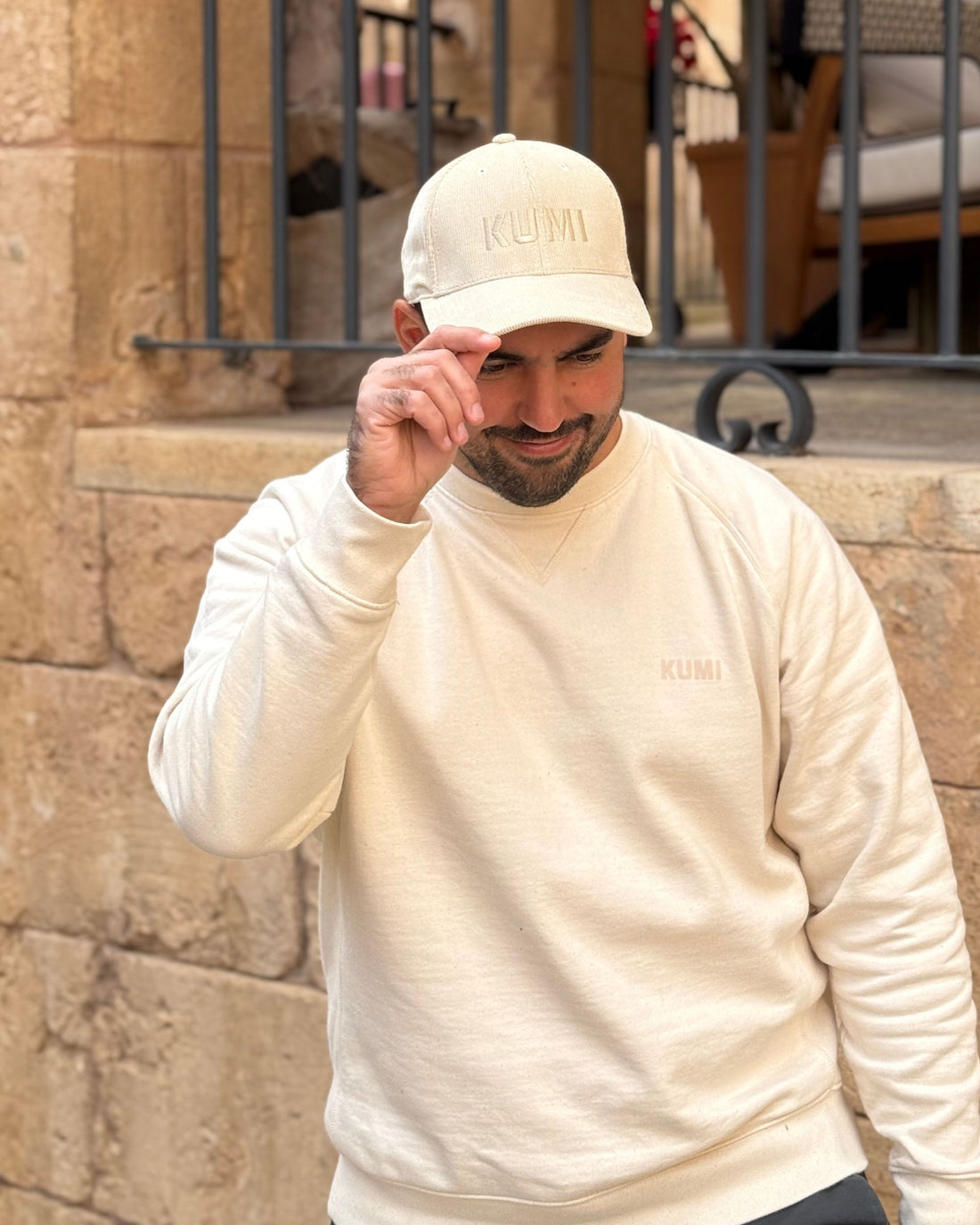 Hombre ajustándose la gorra KUMI color natural, llevando una sudadera a juego, en una calle con paredes de piedra