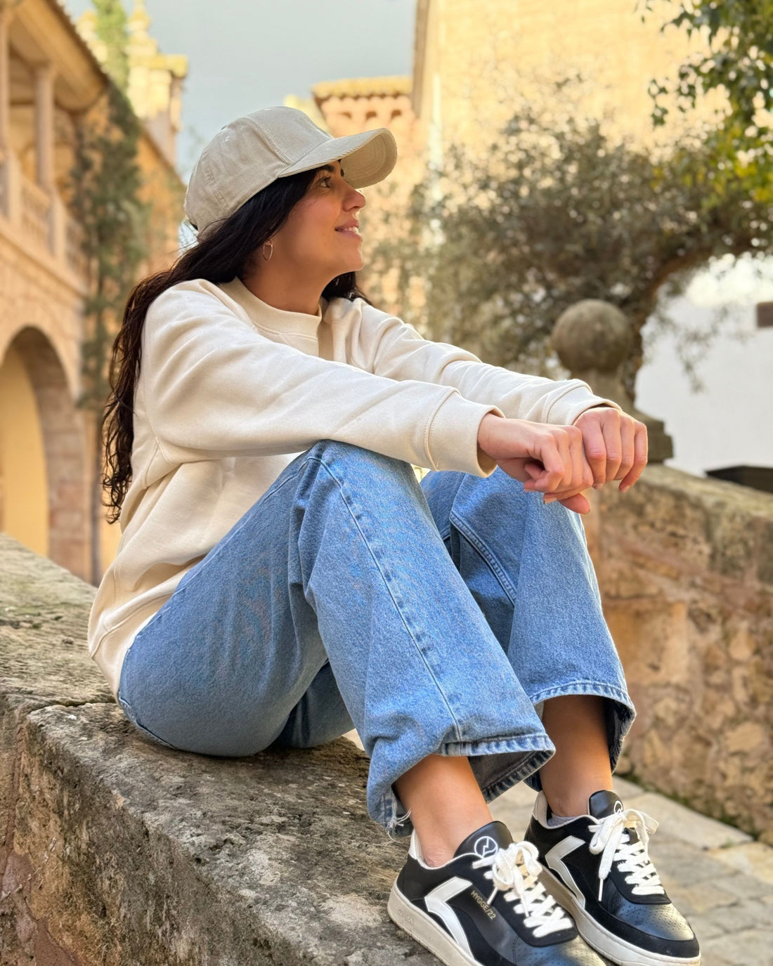 Mujer sentada sobre un muro de piedra en un entorno exterior, vistiendo gorra KUMI color natural, sudadera natural y vaqueros, mirando hacia un lado y sonriendo