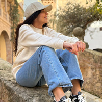 Mujer sentada sobre un muro de piedra en un entorno exterior, vistiendo gorra KUMI color natural, sudadera natural y vaqueros, mirando hacia un lado y sonriendo