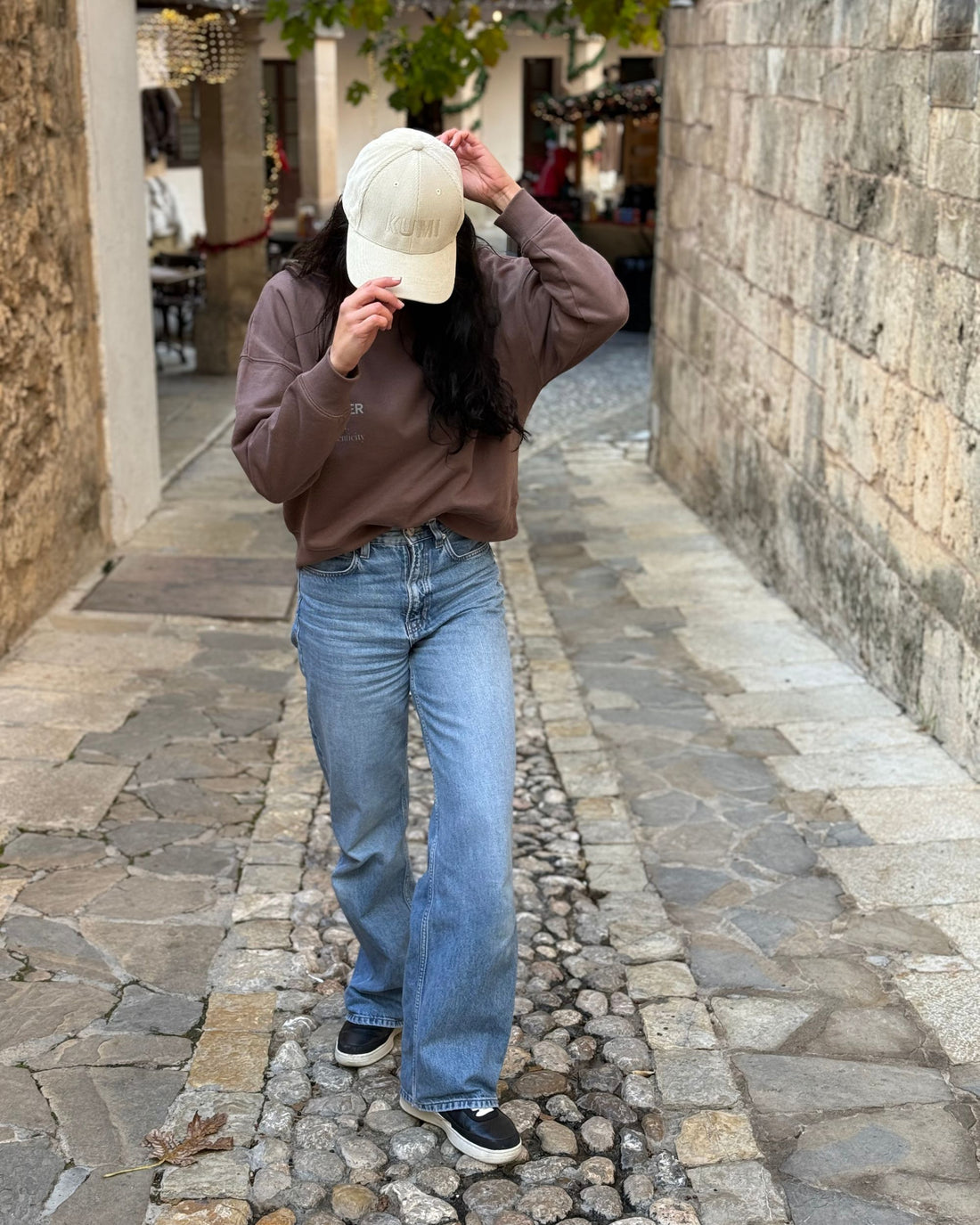 Mujer caminando por una calle empedrada con sudadera color moka y gorra KUMI natural, sujetando la visera mientras mira hacia abajo