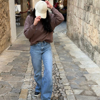Mujer caminando por una calle empedrada con sudadera color moka y gorra KUMI natural, sujetando la visera mientras mira hacia abajo