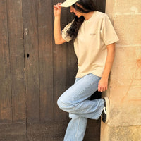 Mujer apoyada en una puerta de madera oscura, vistiendo camiseta y gorra KUMI color natural, con vaqueros y zapatillas negras