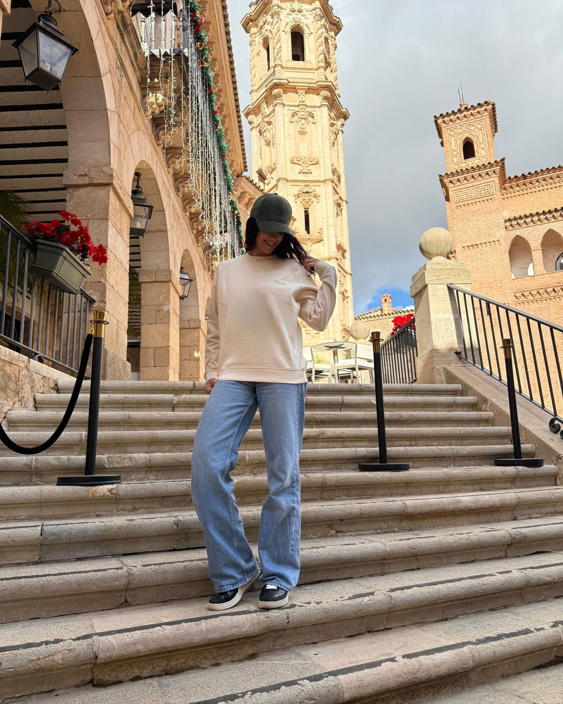 Modelo femenina con la KUMI Natural Unisex Crewneck en color natural, look casual en escaleras con arquitectura mediterránea