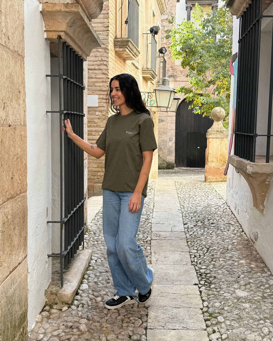 Camiseta KUMI verde oliva de algodón orgánico, corte regular, en look casual caminando por calle empedrada