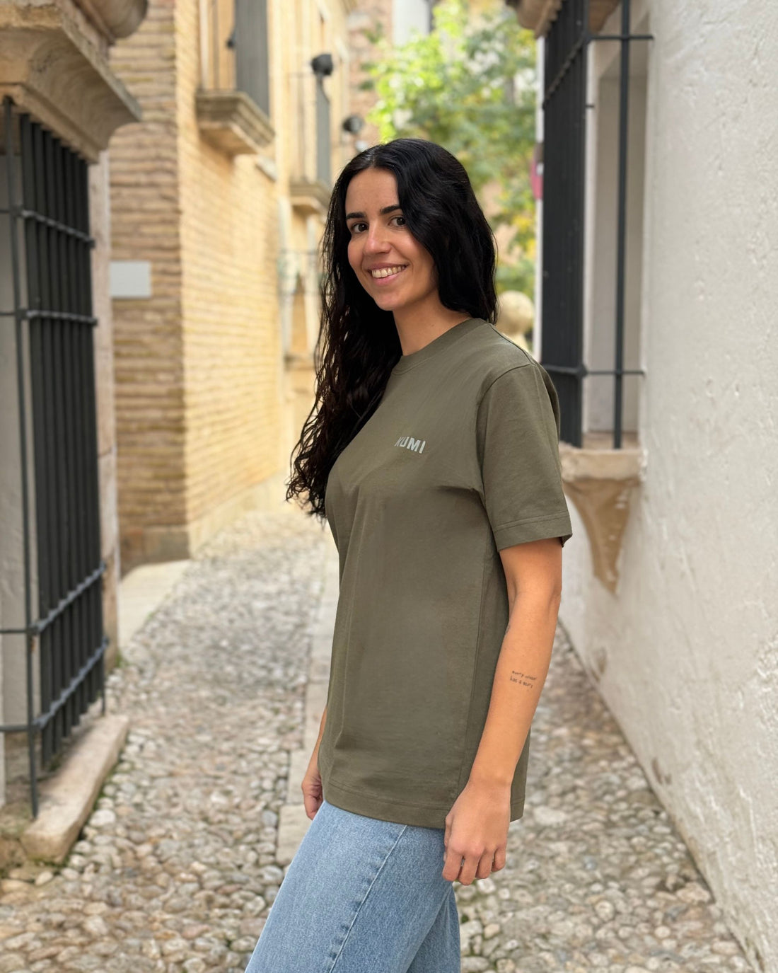 Modelo sonriendo con Camiseta KUMI verde oliva de algodón orgánico, corte regular, en calle empedrada